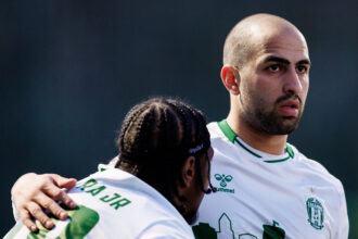 Two soccer players in white and green jerseys share a hug, the foreground player with a shaved head and beard looking off to the right. Two teammates in white and green athletic jerseys stand close, one with cornrows and the other with a shaved head.
