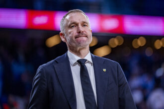 Man in a dark suit and tie with a small lapel pin, standing against a blurred pink-purple light background.