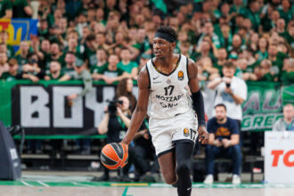 Basketball player in a white jersey dribbles the ball on a court with a cheering green-clad crowd in the background.