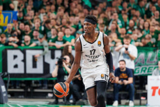 Basketball player in a white jersey dribbles the ball on a court with a cheering green-clad crowd in the background.