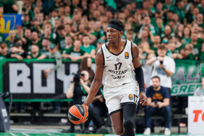 Basketball player in a white jersey dribbles the ball on a court with a cheering green-clad crowd in the background.