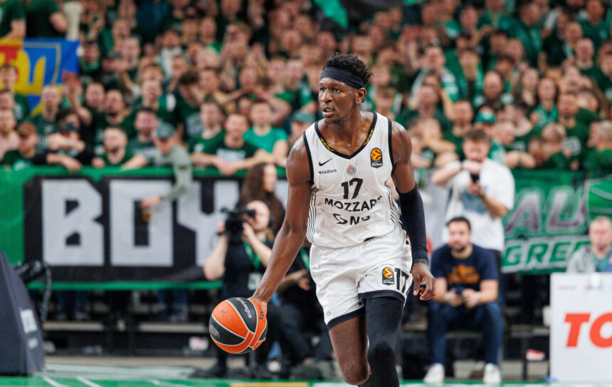 Basketball player in a white jersey dribbles the ball on a court with a cheering green-clad crowd in the background.
