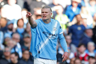 Manchester City footballer in a sky-blue jersey points and celebrates during a match, audience in the background
