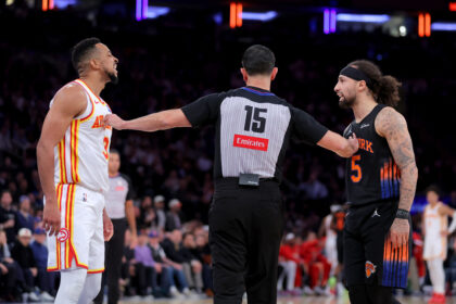 Two basketball players face off as a referee stands between them on the court during a game; left player wears a white jersey with red and yellow trim, right player wears a dark jersey with orange accents and a headband, with a blurred crowd in the background.