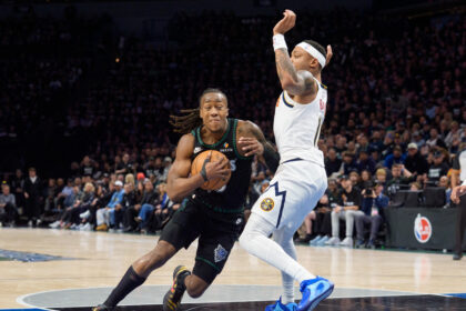 Two basketball players in action competing for the ball during a game, one in black uniform clutching the ball while a defender in white jersey reaches in, arena crowd in background