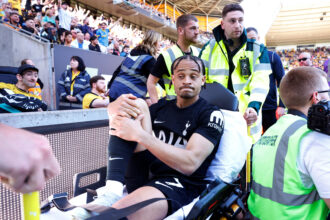 Athlete on a stretcher being carried by staff in high-visibility vests at a stadium, with cheering crowd in the background.