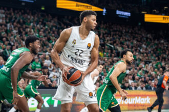 Basketball player in a white Real Madrid jersey (#22) drives with the ball while green-clad defenders surround him in a crowded arena.
