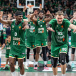 Basketball players in green Zalgiris jerseys raise their arms on court, cheering as fans watch from the stands.