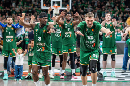 Basketball players in green Zalgiris jerseys raise their arms on court, cheering as fans watch from the stands.