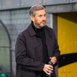 Man in a dark coat holding a plastic bottle, standing outdoors near a yellow wall.