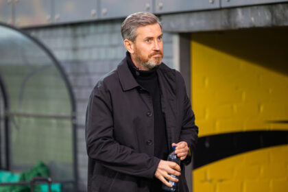 Man in a dark coat holding a plastic bottle, standing outdoors near a yellow wall.