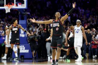 Sixers guard in black jersey (#0) with arms raised in celebration on the court amid cheering fans behind him.