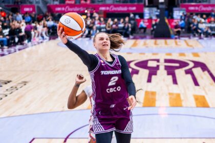 Female basketball players in purple Toronto jerseys jump toward the hoop, reaching for the ball as the crowd watches.