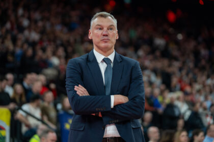 Man in a dark navy suit and tie stands with arms crossed at a crowded arena venue in the background.