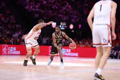Basketball player in a black uniform dribbles toward the basket while defended by a white-uniform opponent in a crowded arena with red advertising boards behind them.