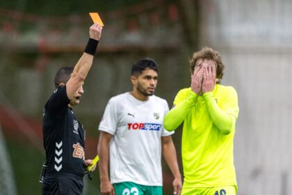 Soccer referee raises an orange card as two players watch—the white-shirted player and a neon-yellow goalkeeper covering his face in the background.