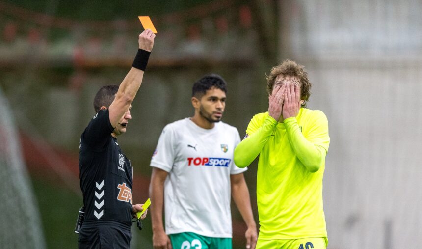 Soccer referee raises an orange card as two players watch—the white-shirted player and a neon-yellow goalkeeper covering his face in the background.