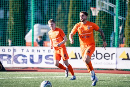 Two soccer players in orange uniforms sprint on a green turf with a ball at their feet, a goal net in the background.