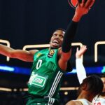 Basketball player in green jersey #0 leaps toward the hoop for a shot while defenders reach up nearby.