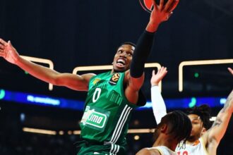 Basketball player in green jersey #0 leaps toward the hoop for a shot while defenders reach up nearby.