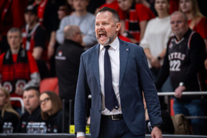 Man in a blue suit and tie shouting on the sidelines during a game, crowd in red behind him.