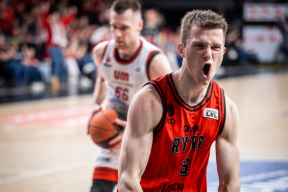 Basketball player in red jersey screams with mouth wide open during a fast-paced game, arena crowd blurred in background