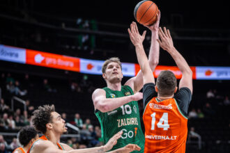 Two basketball players in green and orange battle for the ball in mid-air as hands reach up during a play, with a packed arena in the background.