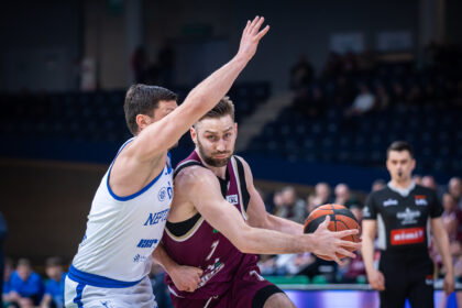 Two basketball players clash for the ball, one in white with blue trim reaching up while the other in maroon guards the ball in a crowded arena.