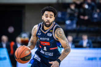 Basketball player in a blue jersey dribbles an orange ball, focused on the game with visible tattoos on both arms.