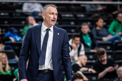 Man in a navy suit and dark tie walking courtside in a sports arena, with blurred spectators in the background.