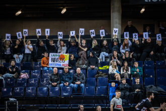 Crowd of spectators in a sports arena holding up letter cards to spell a message across the stands. The signs form a long row above the crowd.