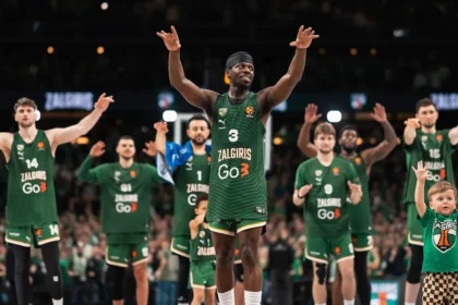 Zalgiris basketball players in green uniforms raise their arms in celebration on the court, with a child in a matching shirt on the right.