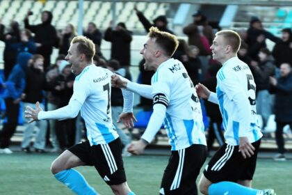 Soccer players in light blue jerseys celebrate a goal on a green field with cheering fans in the stands behind themside.