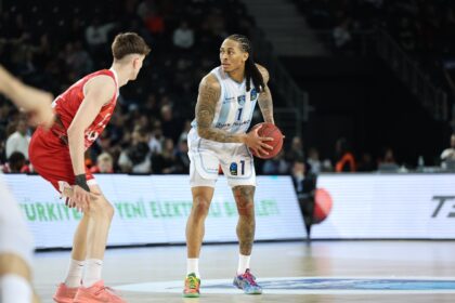 Basketball player with braided hair in a white/blue uniform holding the ball, guarded by a red-uniform defender on a brightly lit court.