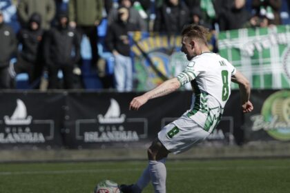 Soccer player in white-green kit (number 8) kicks the ball on a field with spectators in the stands behind him.