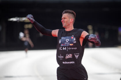 Male athlete raises fists in victory, wearing black sleeveless race kit with bib number 9 and sponsor logos, on an ice rink.