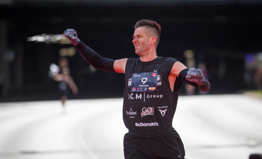 Male athlete raises fists in victory, wearing black sleeveless race kit with bib number 9 and sponsor logos, on an ice rink.