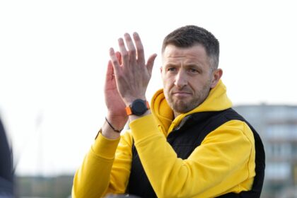 Man in a yellow hoodie and black vest clapping his hands outdoors, with a focused expression.