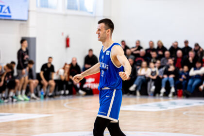 Male basketball player in a blue jersey walking across a court, fists clenched in celebration, with a crowd of spectators watching from the stands in the background.