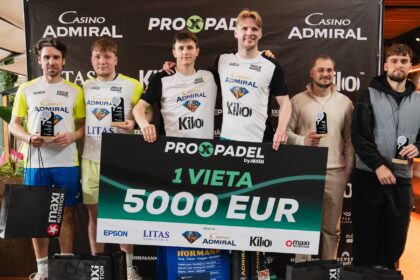 Six competitors stand on a podium holding trophies and a large 5,000 EUR prize check, with sponsor banners in the background and black gift bags at their feet.