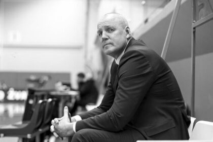 Man in a dark suit sits on a bench in an indoor gym or arena, looking toward the camera.