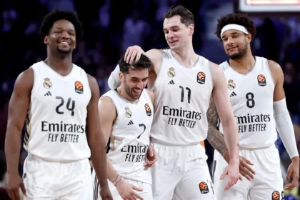 Four Real Madrid basketball players in white jerseys celebrate on the court, with one patting a teammate on the head.