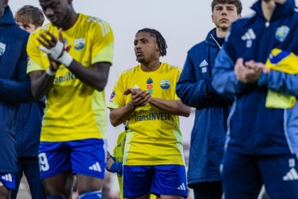 Soccer players in yellow jerseys clap before a game, central player with braids and tattoos standing out amid teammates in blue jackets.