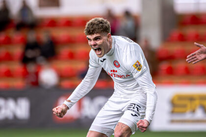 Soccer player in a white jersey celebrates with arms outstretched on a blurred stadium background.