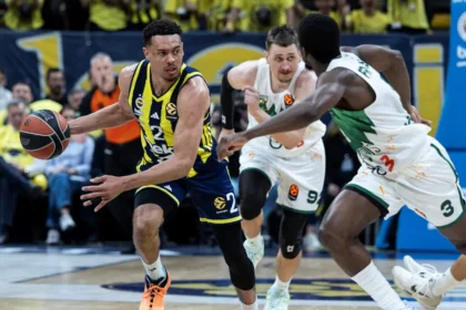 Action shot of a basketball game: player in yellow-blue uniform dribbles past a defender in white-green, with a cheering crowd in the background.
