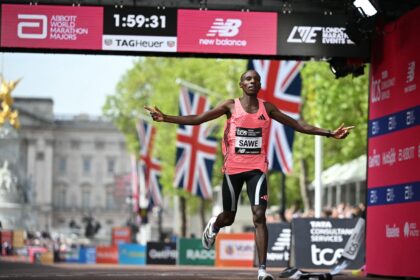 Male marathon runner in a pink sleeveless top crosses the finish line with arms outstretched under sponsor banners and flags in the background.