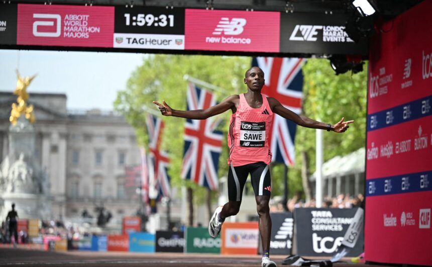 Male marathon runner in a pink sleeveless top crosses the finish line with arms outstretched under sponsor banners and flags in the background.
