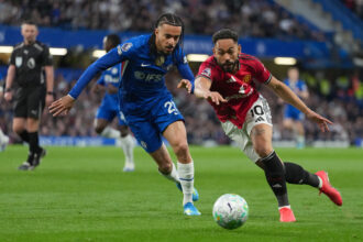 Two soccer players chase the ball on a green pitch; a blue Chelsea player and a red Manchester United player race toward the ball in a crowded stadium