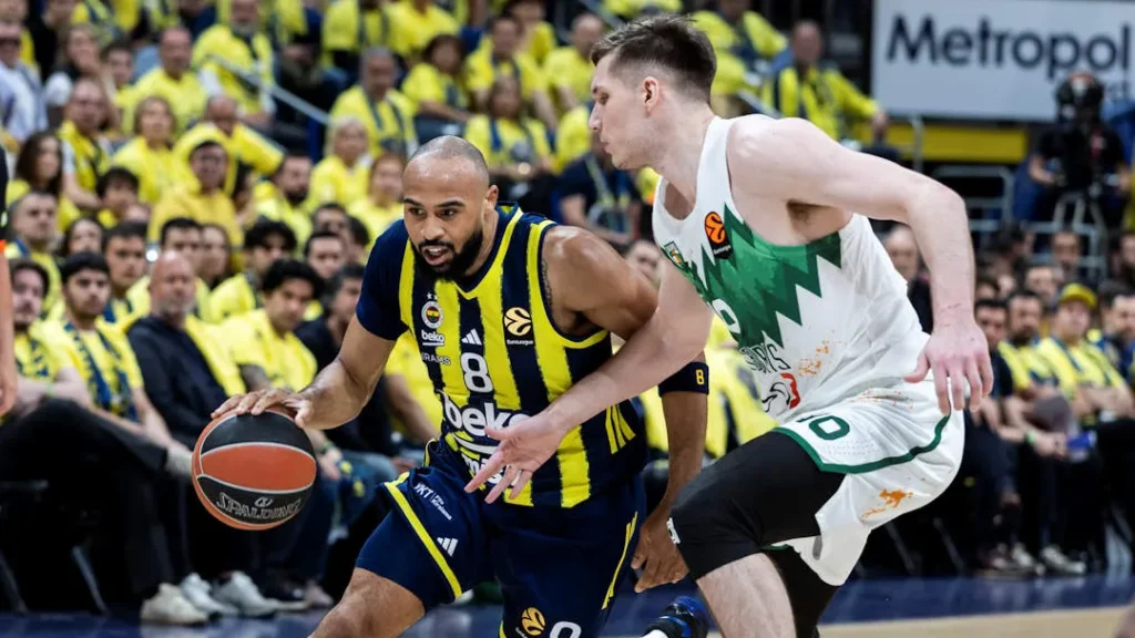 Basketball player in blue-yellow jersey dribbles past defender in white-green jersey during a crowded indoor game.