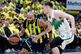 Basketball player in blue-yellow jersey dribbles past defender in white-green jersey during a crowded indoor game.